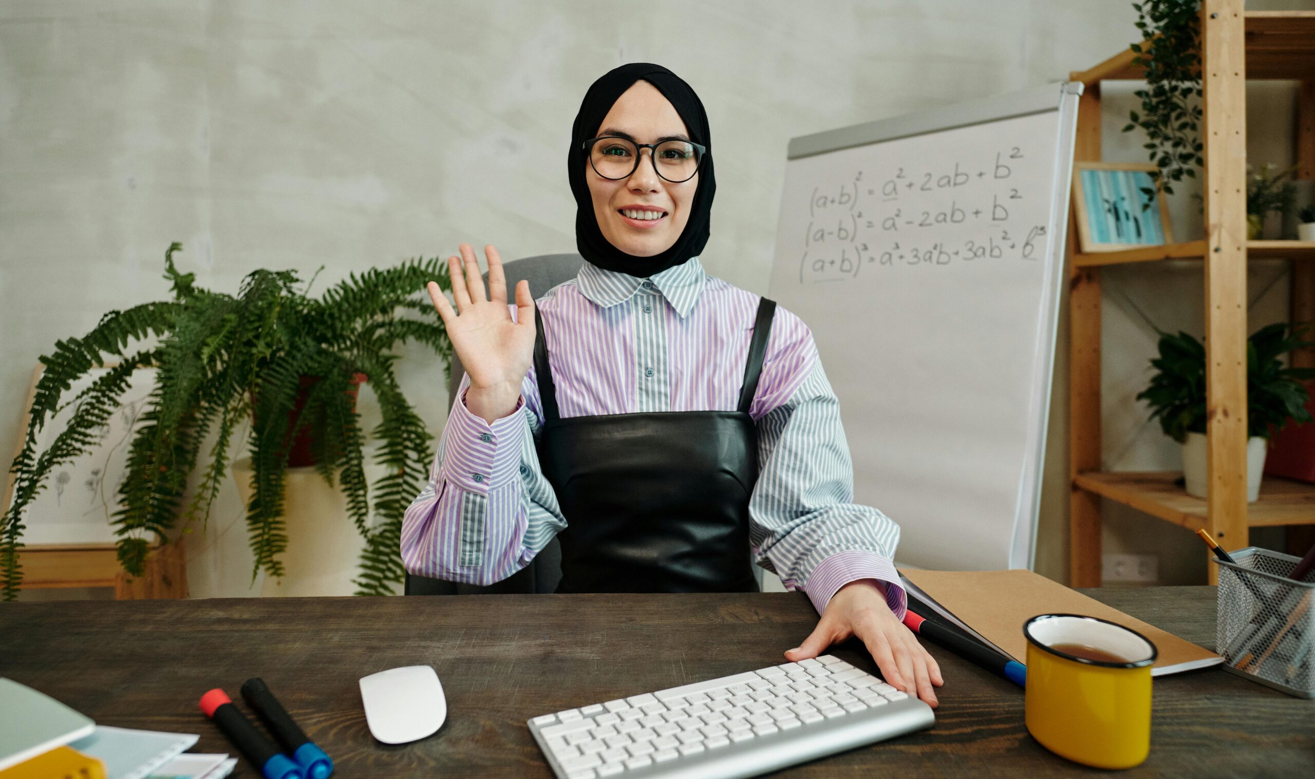Smiling woman in hijab waving at the camera in a modern office setting.